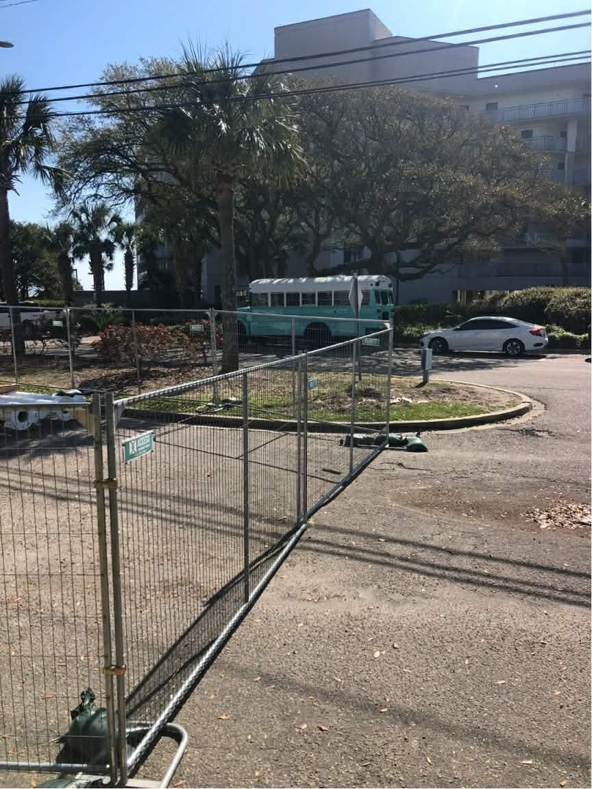 Temporary metal fence in a parking lot beside trees and a large building, with cars in the background