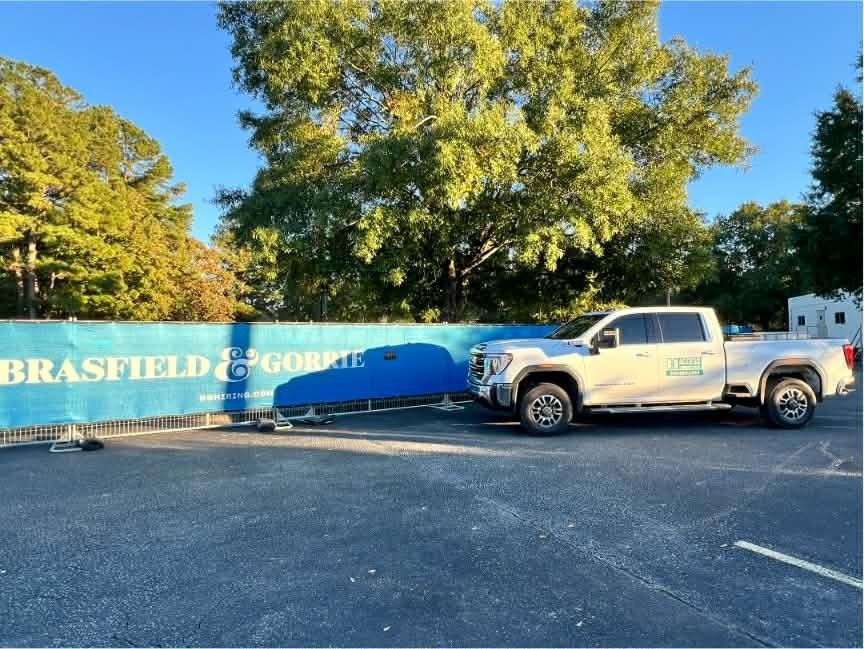 White pickup truck parked beside a blue Brasfield & Gorrie construction sign in a lot under trees