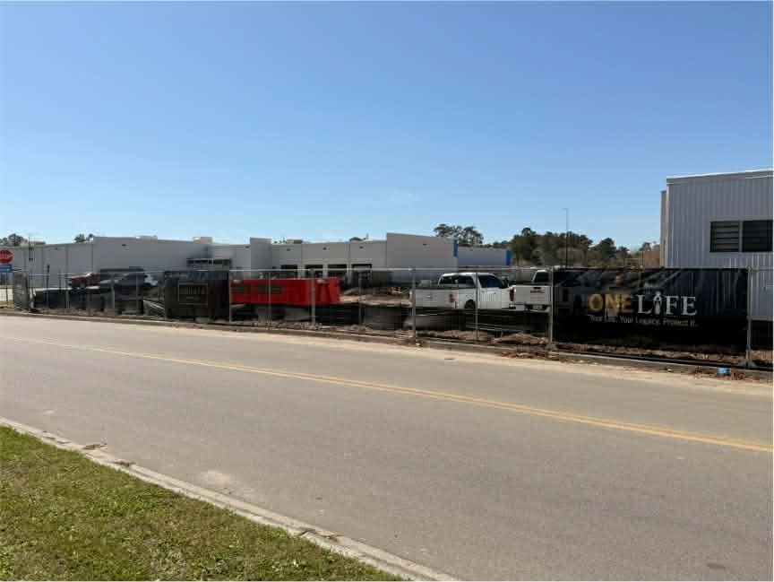 Street view of a fenced lot with parked trucks and a OneLife sign beside a road under a clear blue sky