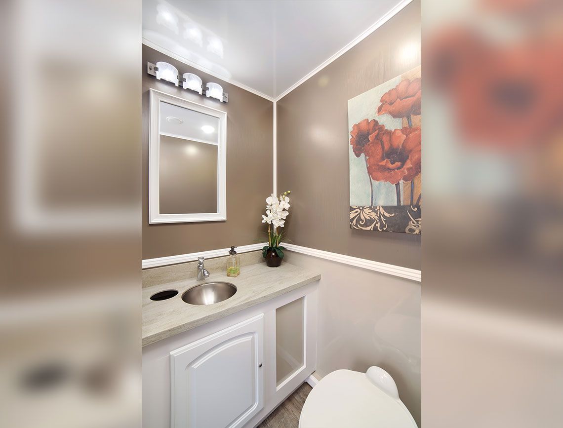Small bathroom vanity with sink, mirror, beige walls, floral wall art, and vase of white flowers.