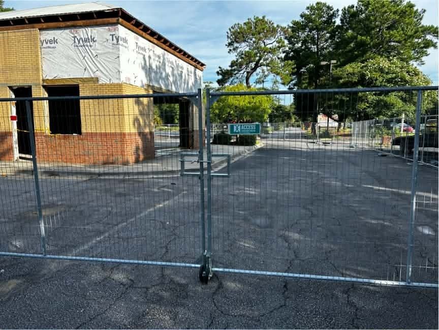 Temporary metal fence blocking an empty paved lot beside a brick building and trees