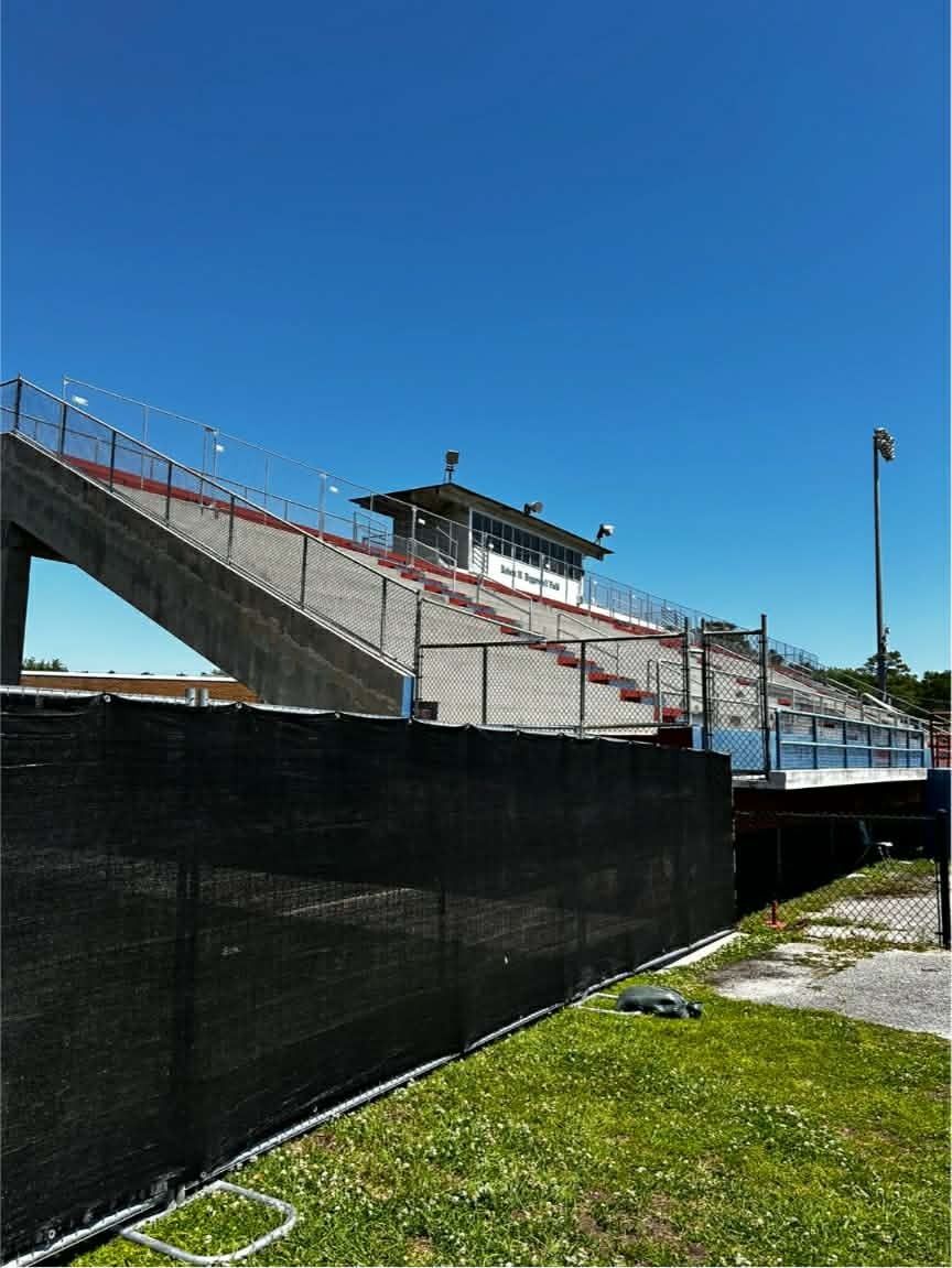 Empty outdoor stadium bleachers under a clear blue sky, viewed from below behind a black fence.