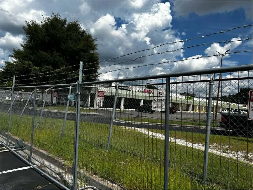 Chain-link fence beside a grassy lot and low buildings under a cloudy sky.
