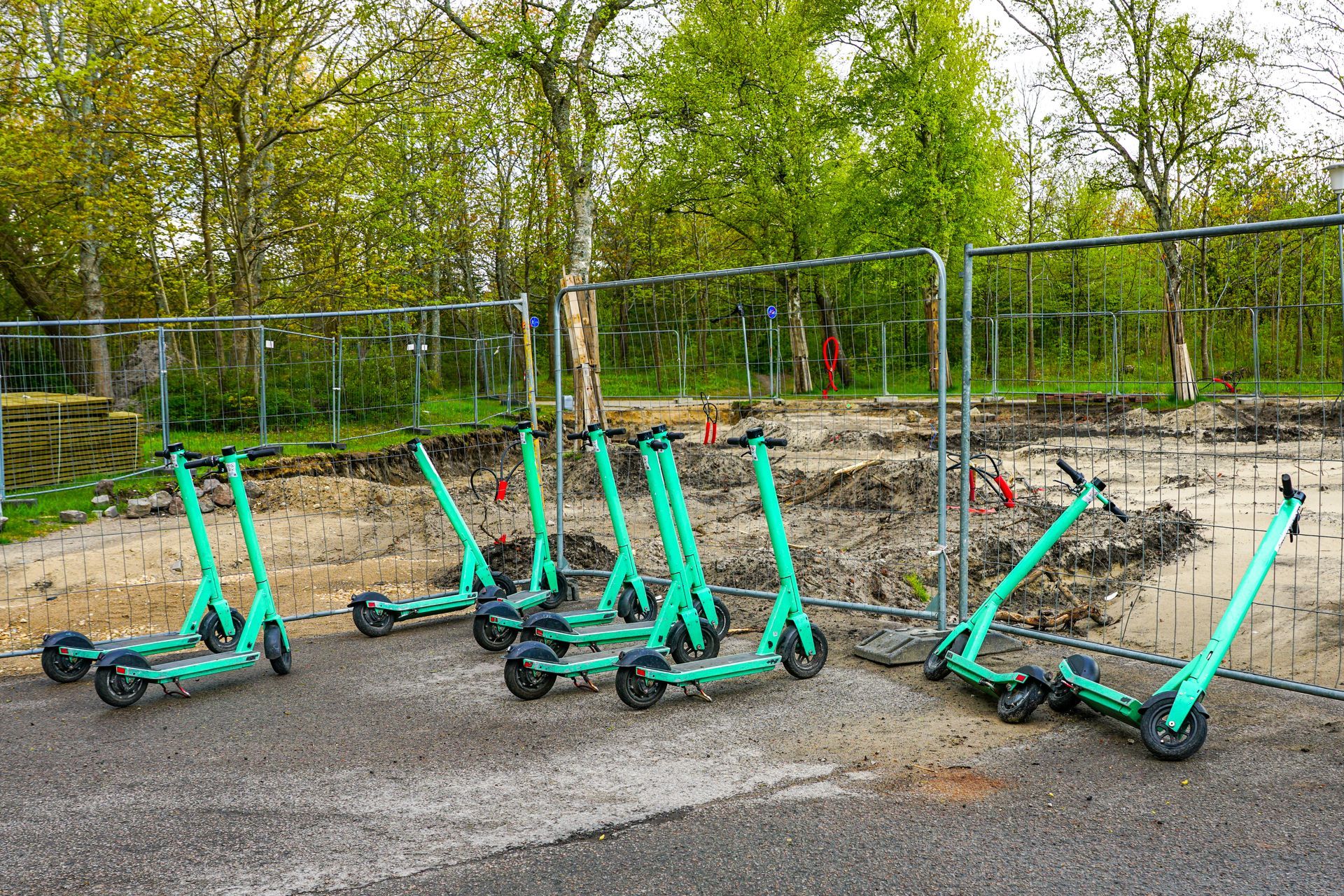 Six green electric scooters parked near a construction site, behind a chain-link fence. Six green electric scooters parked near a construction site, behind a chain-link fence.