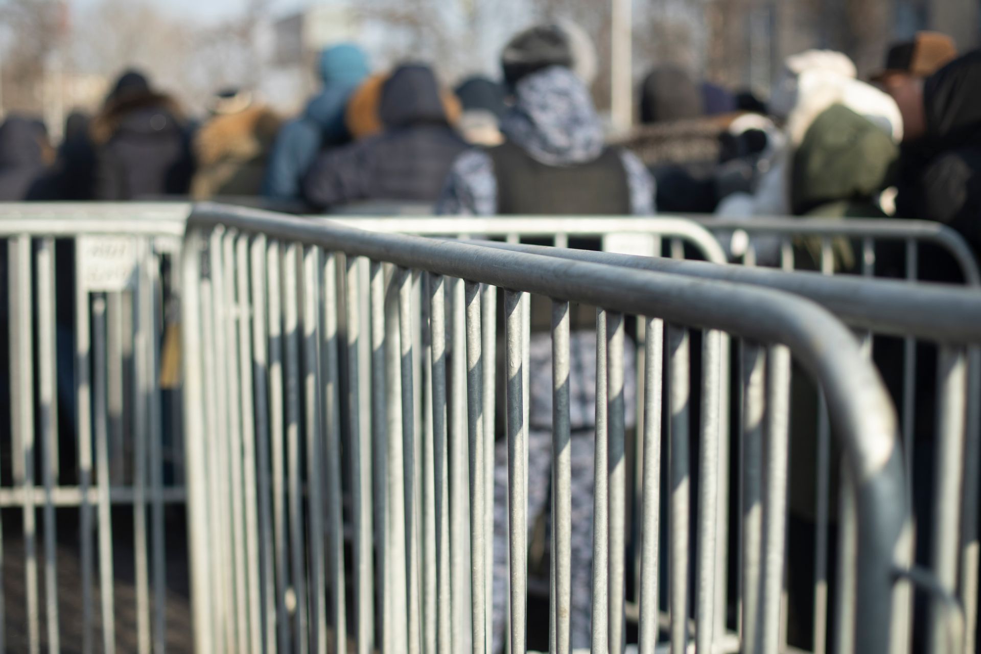 Row of townhouses behind temporary fencing along a sidewalk. Gray and white buildings, overcast sky. Metal barricade with a crowd of people in the blurred background.