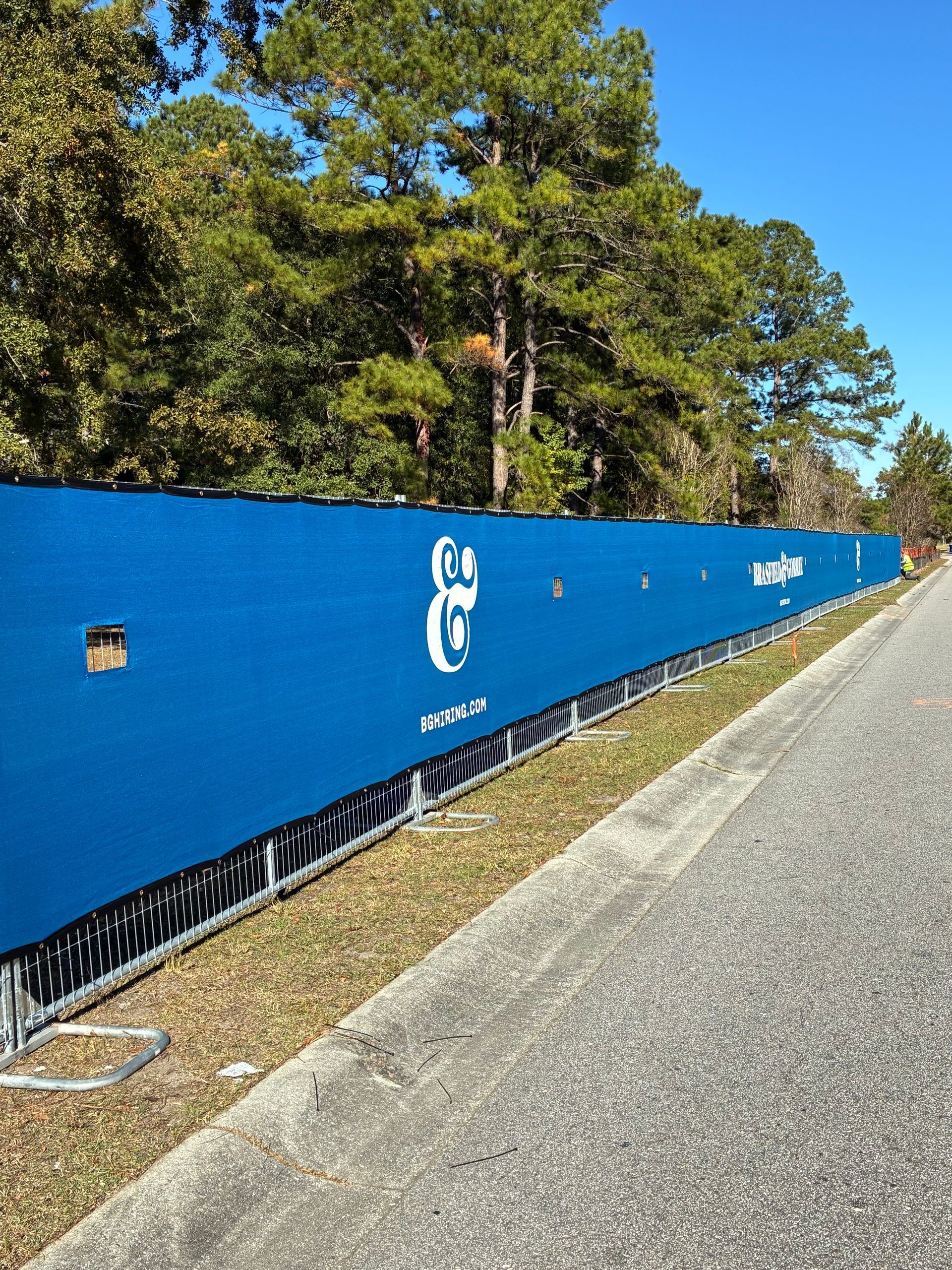 Blue construction fence with logos, alongside a paved pathway and trees.