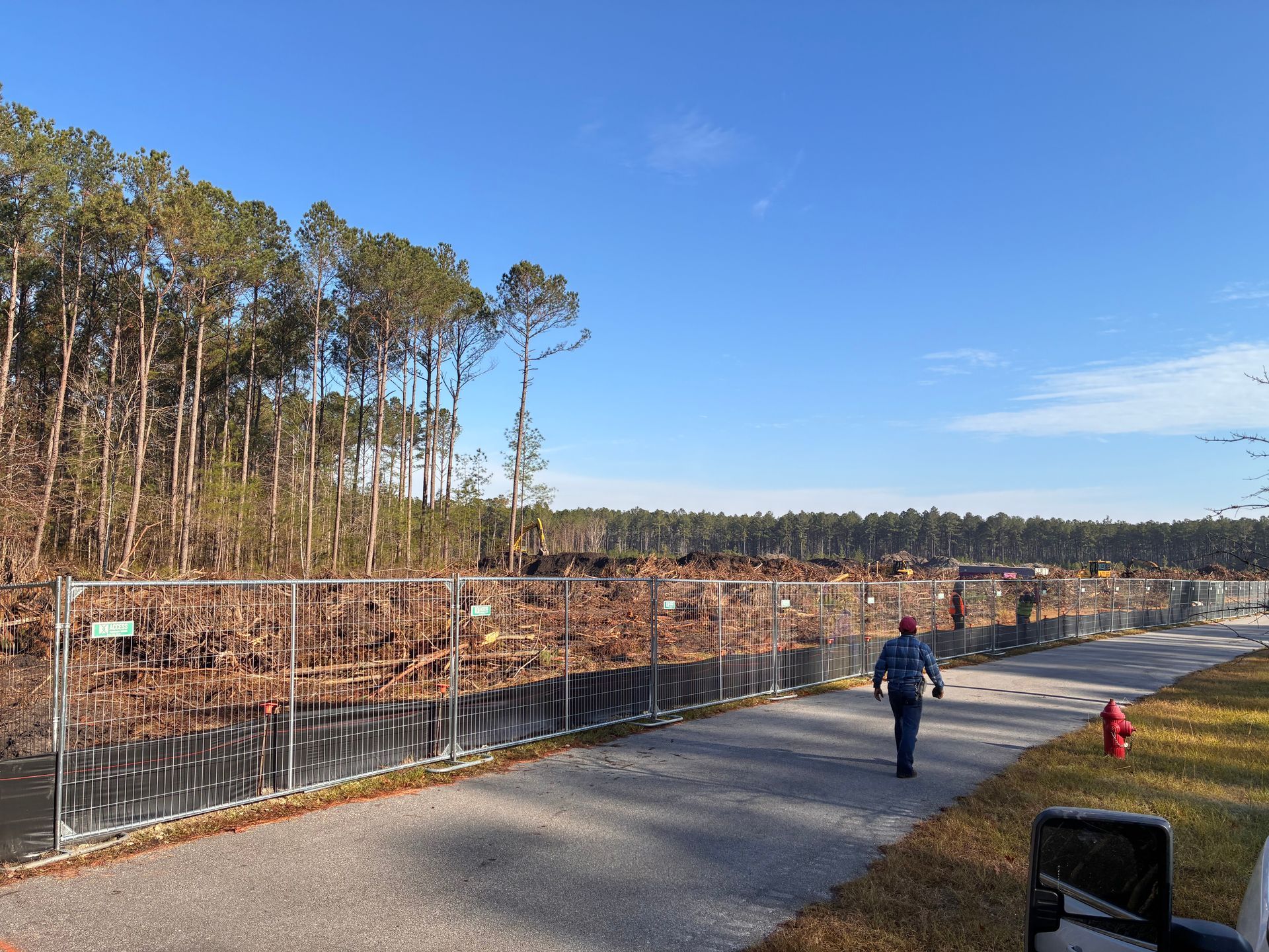 Person walking on a paved path beside a fenced wooded area on a sunny day