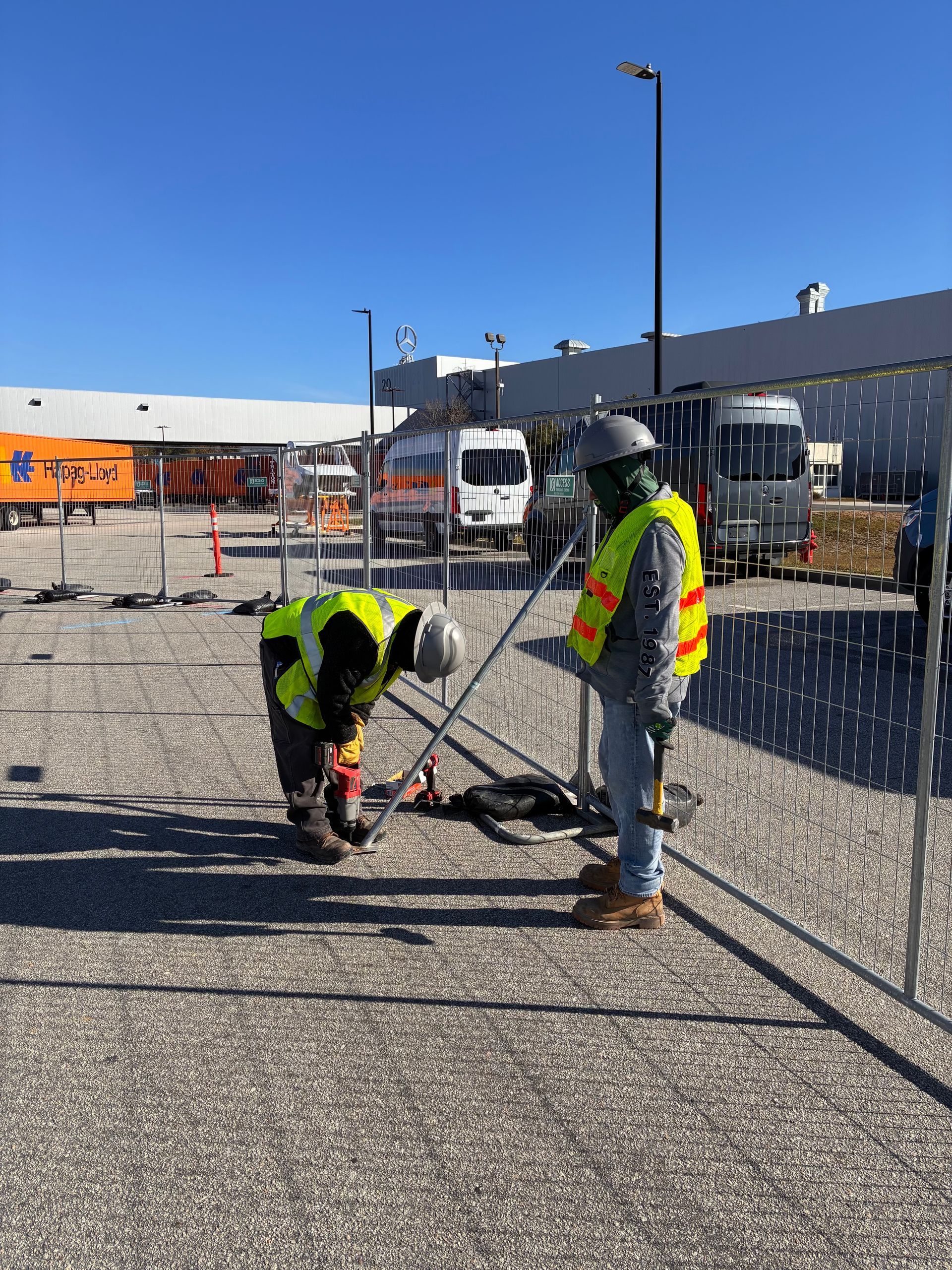 Two workers in safety vests install fencing at a construction site under a clear blue sky