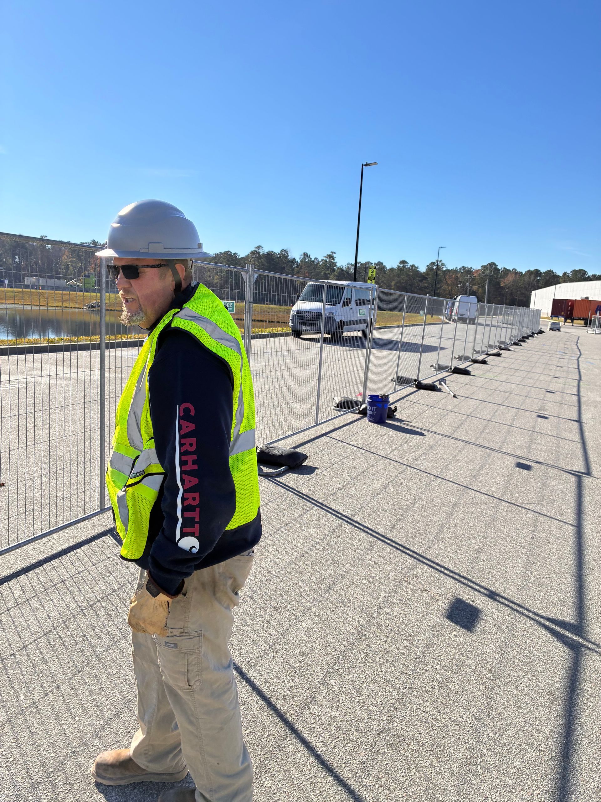 Worker in hard hat and hi-vis vest stands on a gravel rooftop beside safety barriers and equipment.