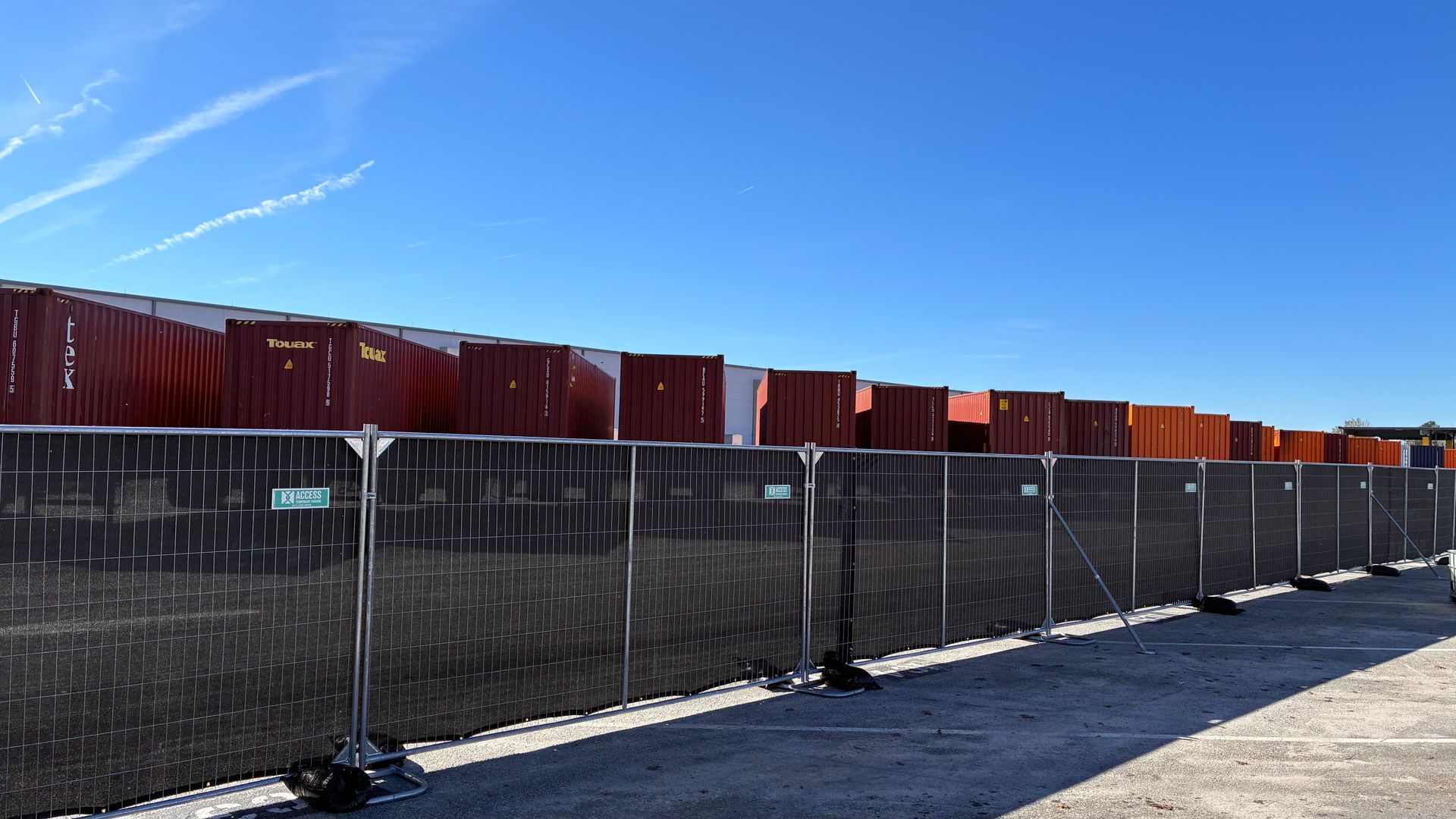 Long red freight containers behind a black chain-link fence under a clear blue sky