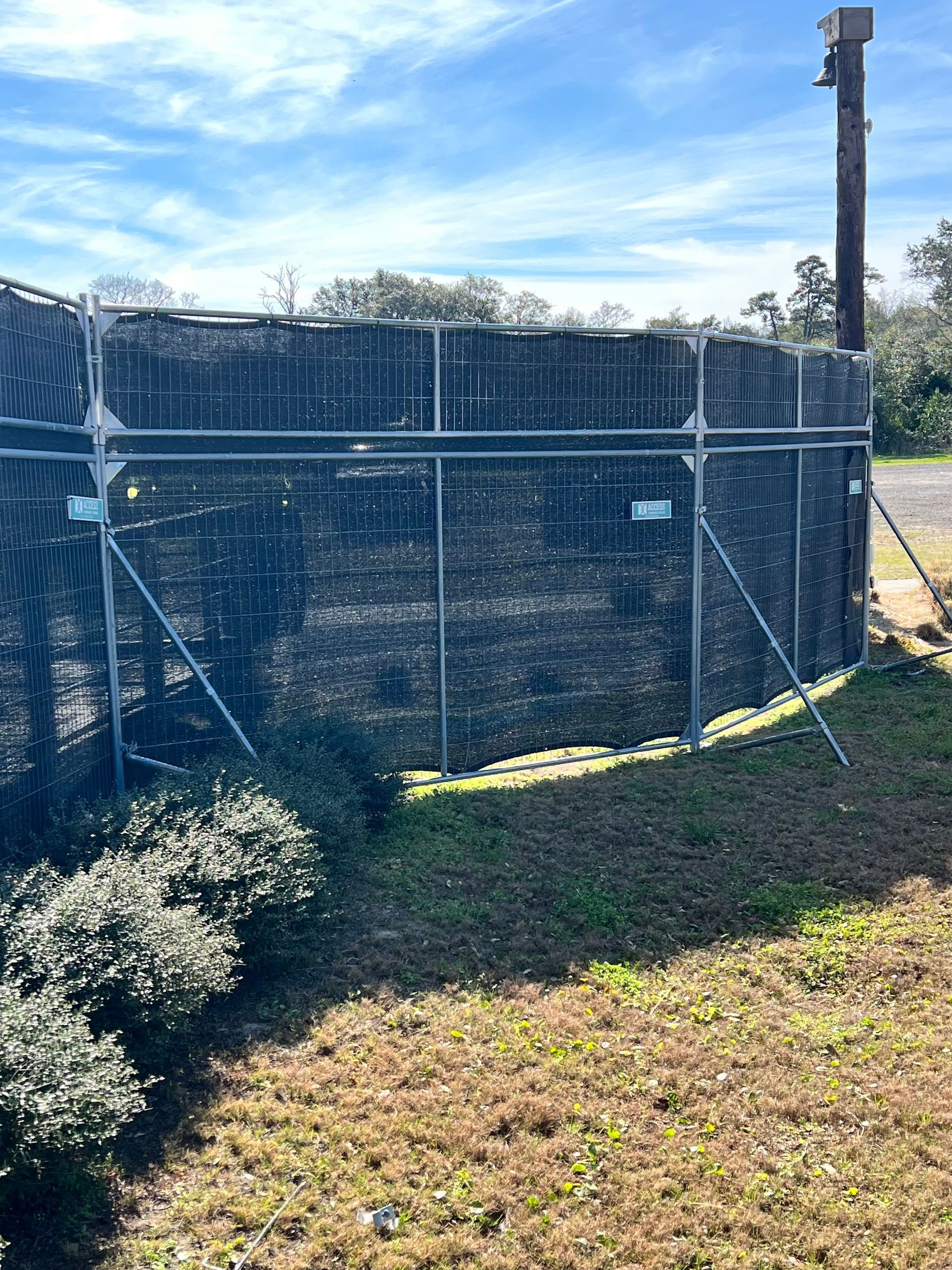 Chain-link fence around a grassy field under a blue sky, with a tall pole and shrubs in the foreground.
