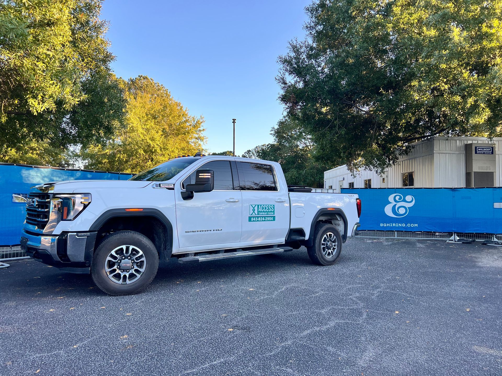 White pickup truck parked beside a blue fenced construction site under trees.