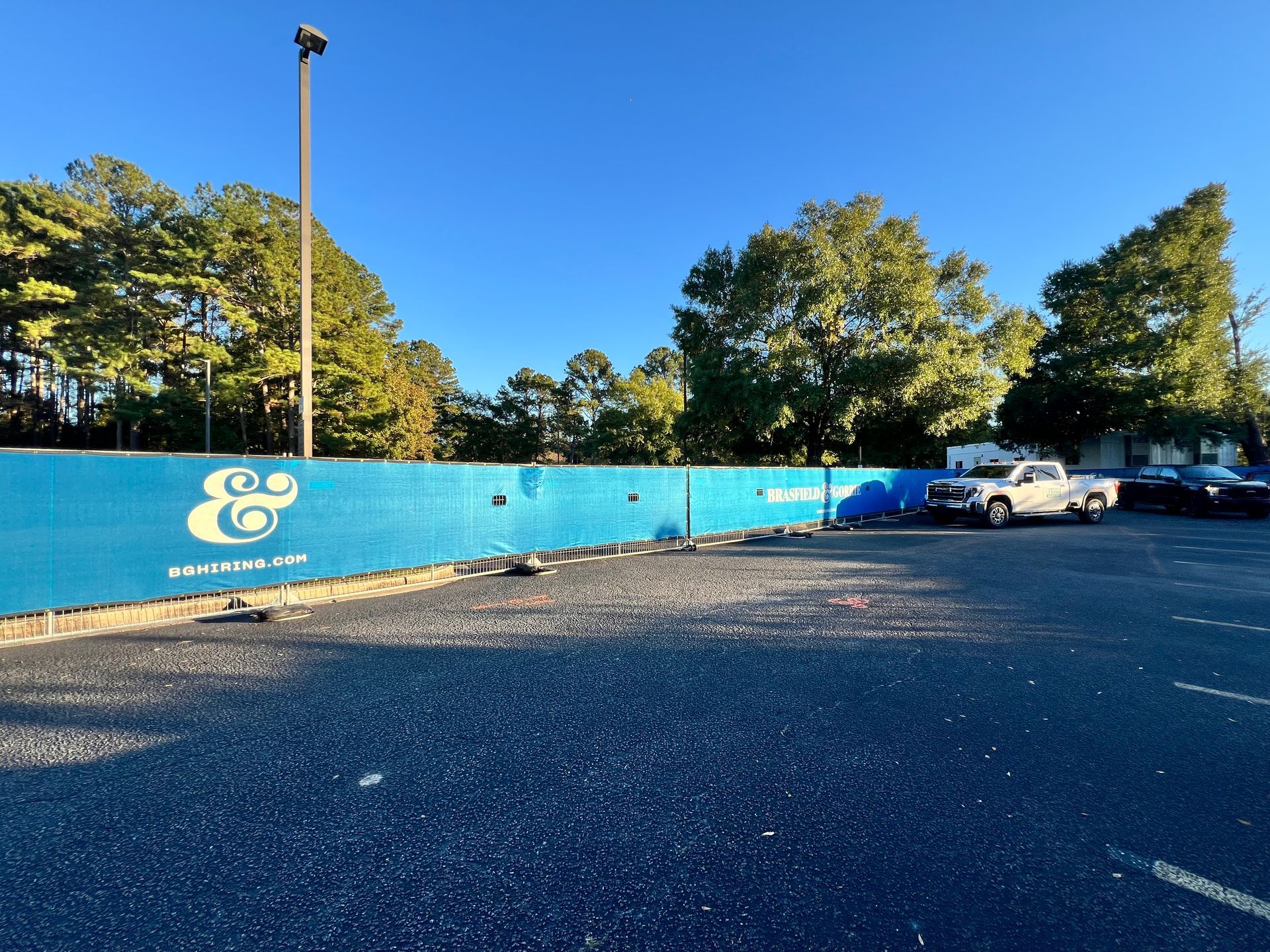 Blue fence along a parking lot with trees behind it, a white pickup truck, and a clear blue sky.
