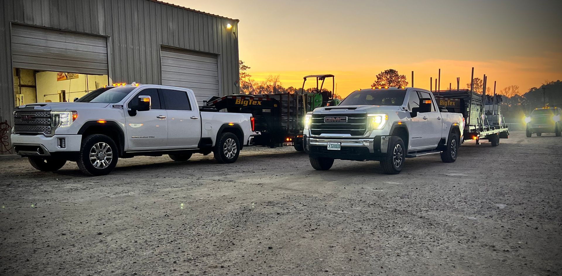 Two white pickup trucks with headlights on parked at dusk beside an industrial building