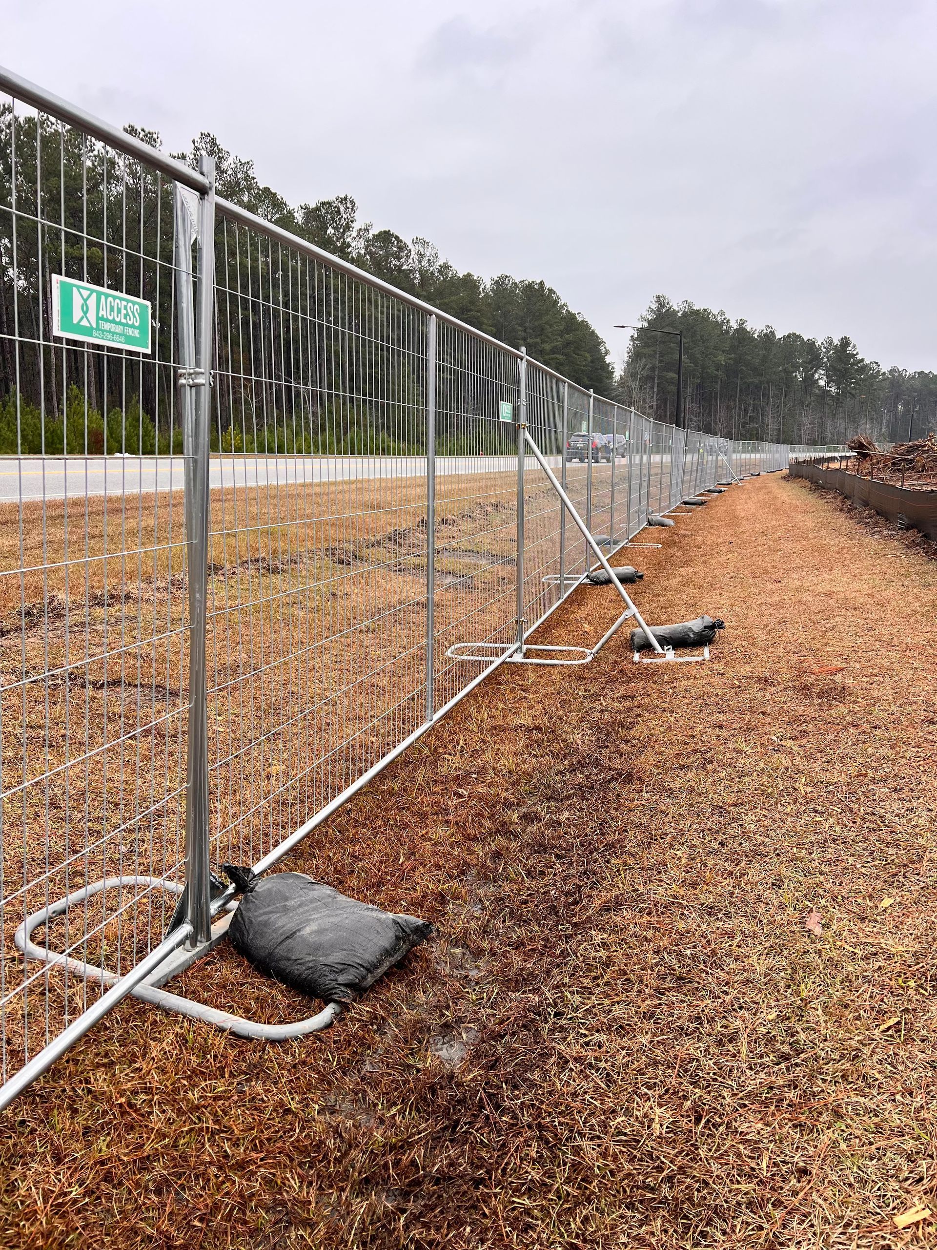 Metal fence along a road beside a pile of wood chips, with trees in the background.