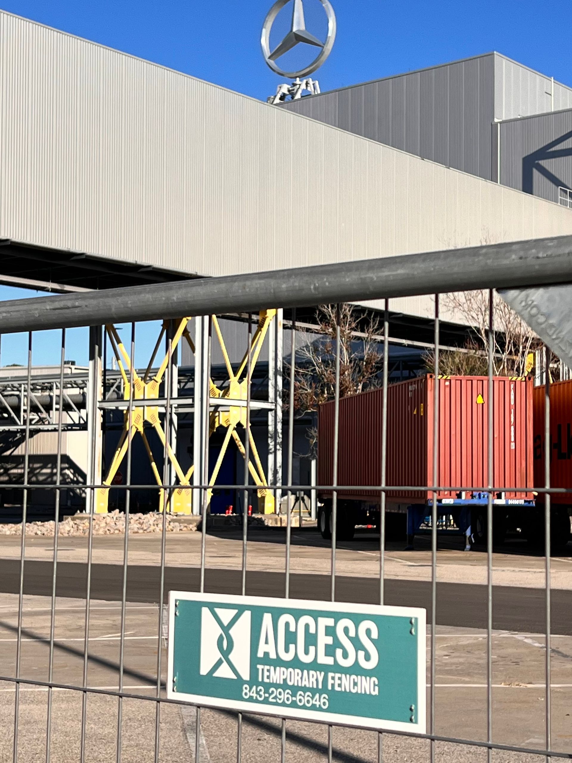 Security gate with metal fence, Access Temporary Fencing sign, and red industrial containers under a Mercedes logo.