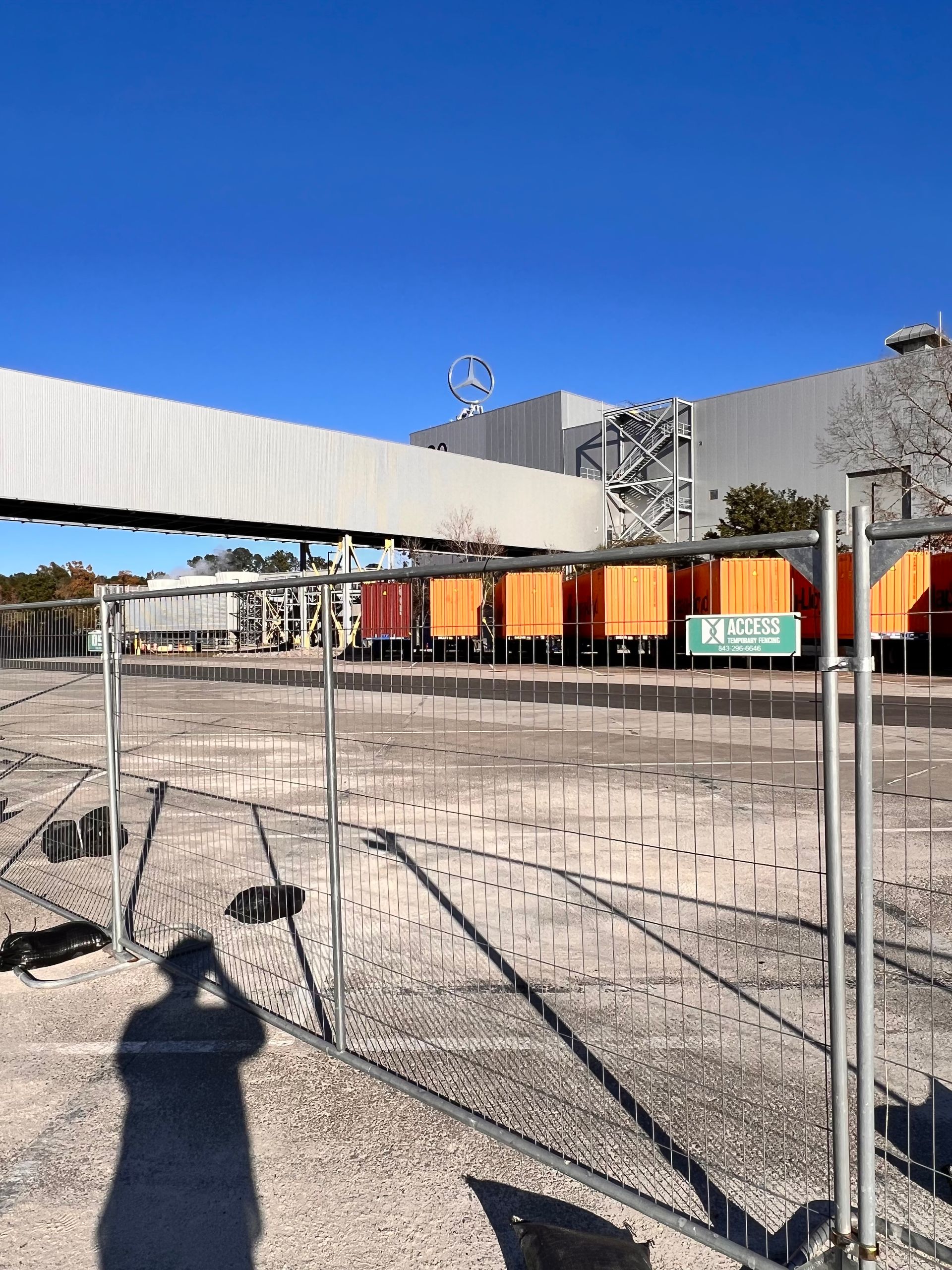 Outdoor construction site with orange barriers, fencing, and a large industrial building under a clear blue sky