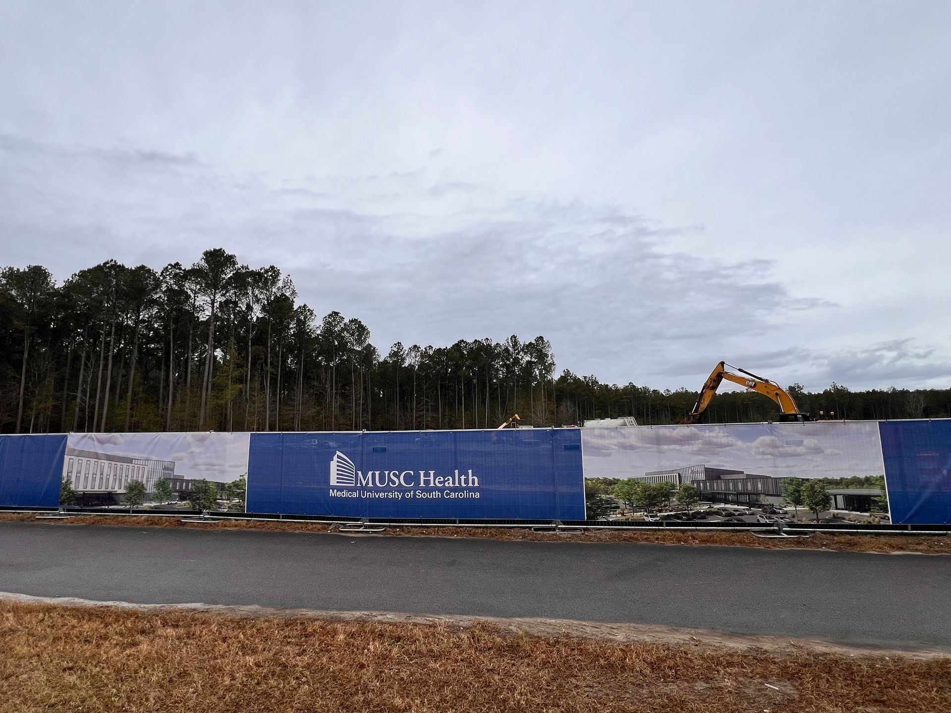 Construction site behind a blue fence with a yellow excavator and overcast sky