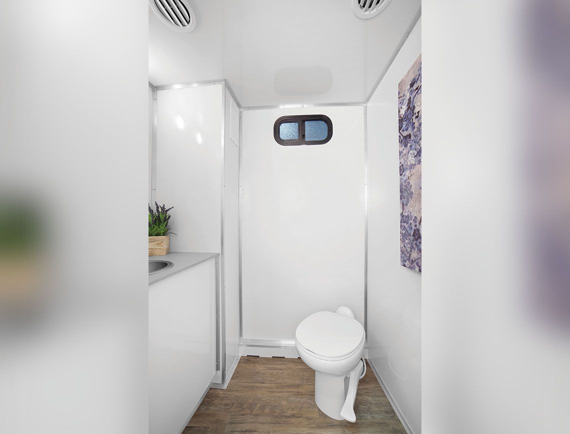 Compact white bathroom with a toilet and small window, narrow wood floor, and a counter with a plant.