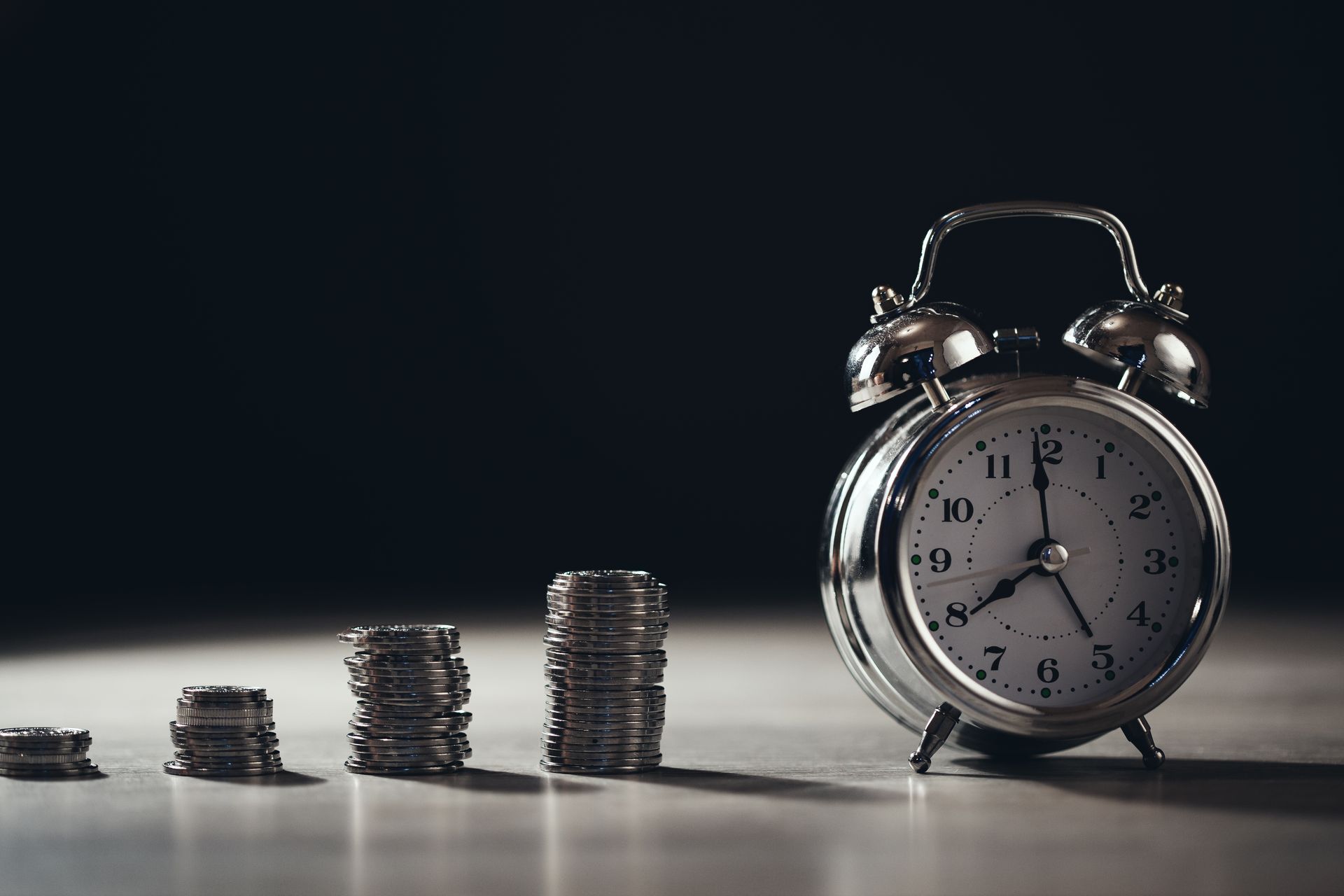 Stacks of coins growing larger next to a silver alarm clock, against a dark background.