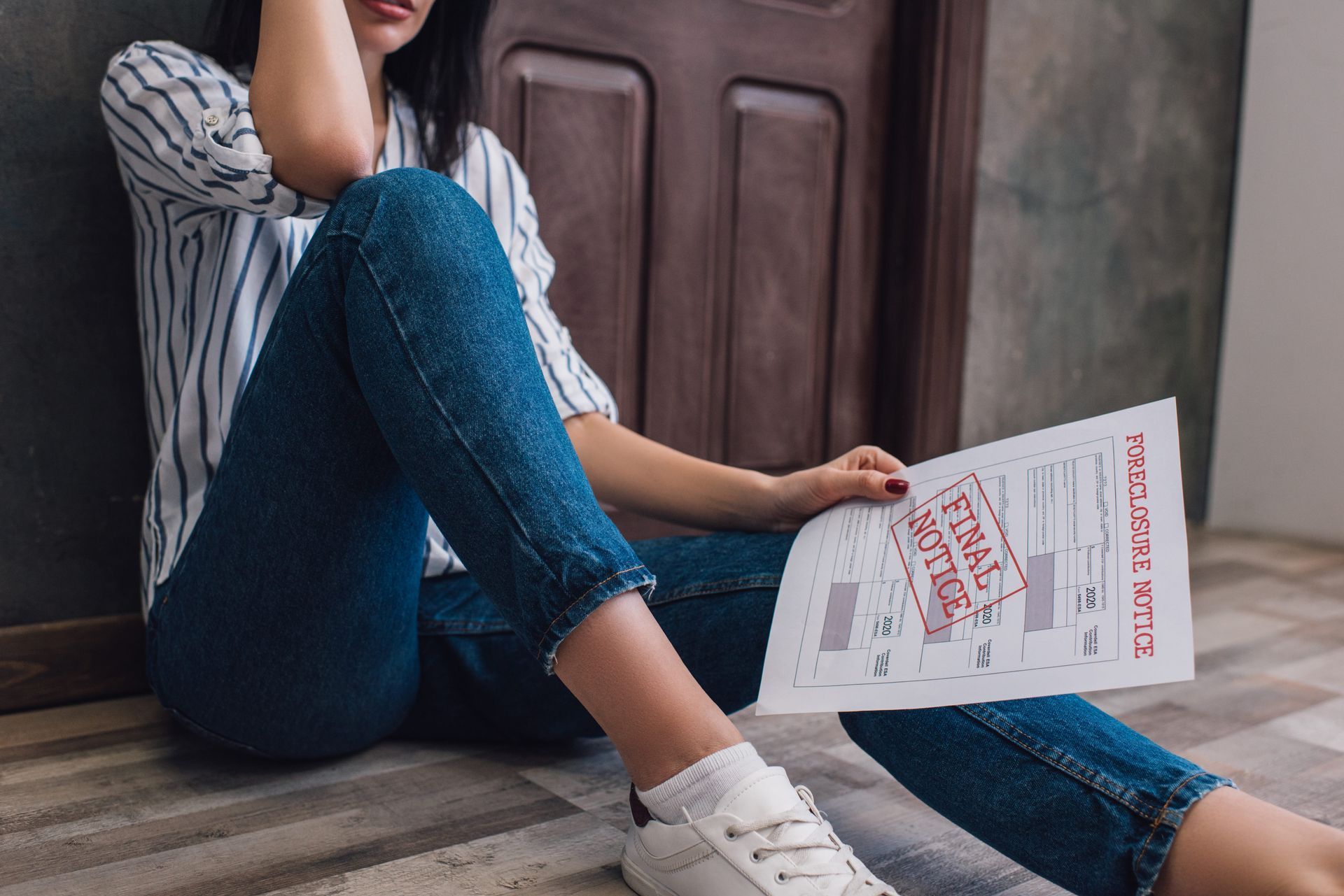 Person sitting by a door, holding a magazine and wearing a striped shirt, blue jeans, and white sneakers.