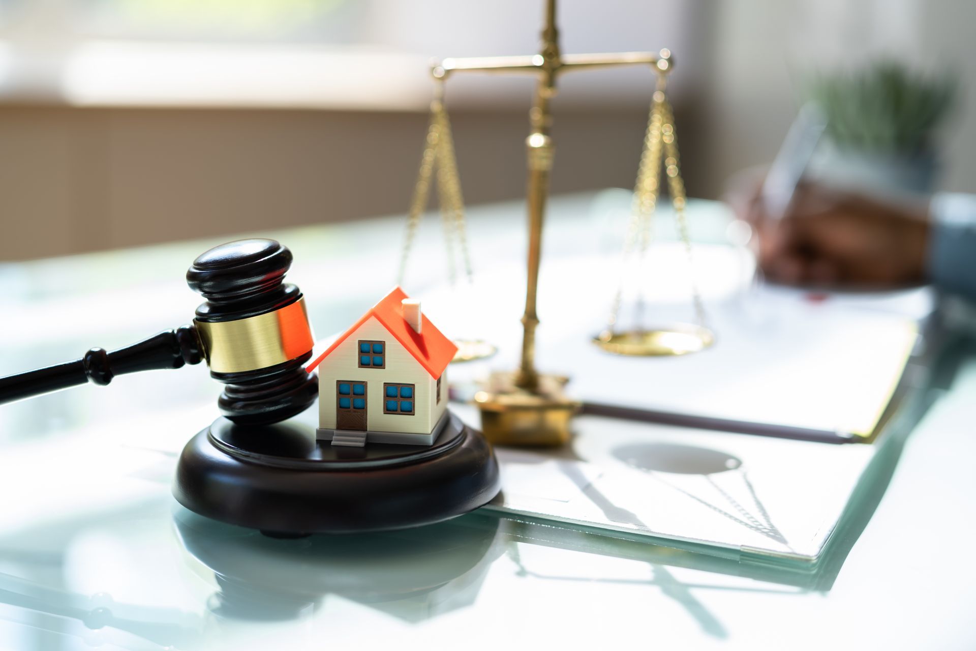 Gavel, toy house, and scales of justice on a desk with a person writing.