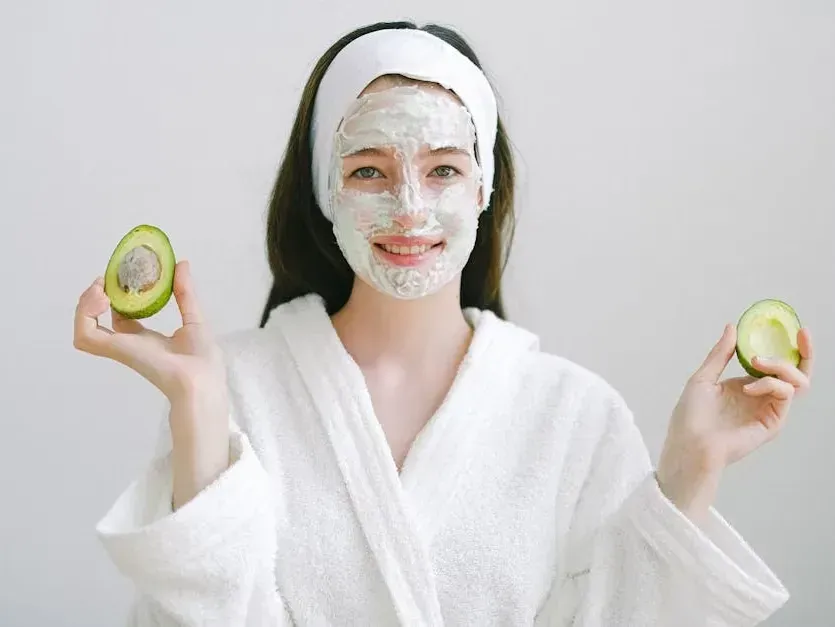 Person in a white robe holding avocado halves, wearing a white facial mask against a plain background