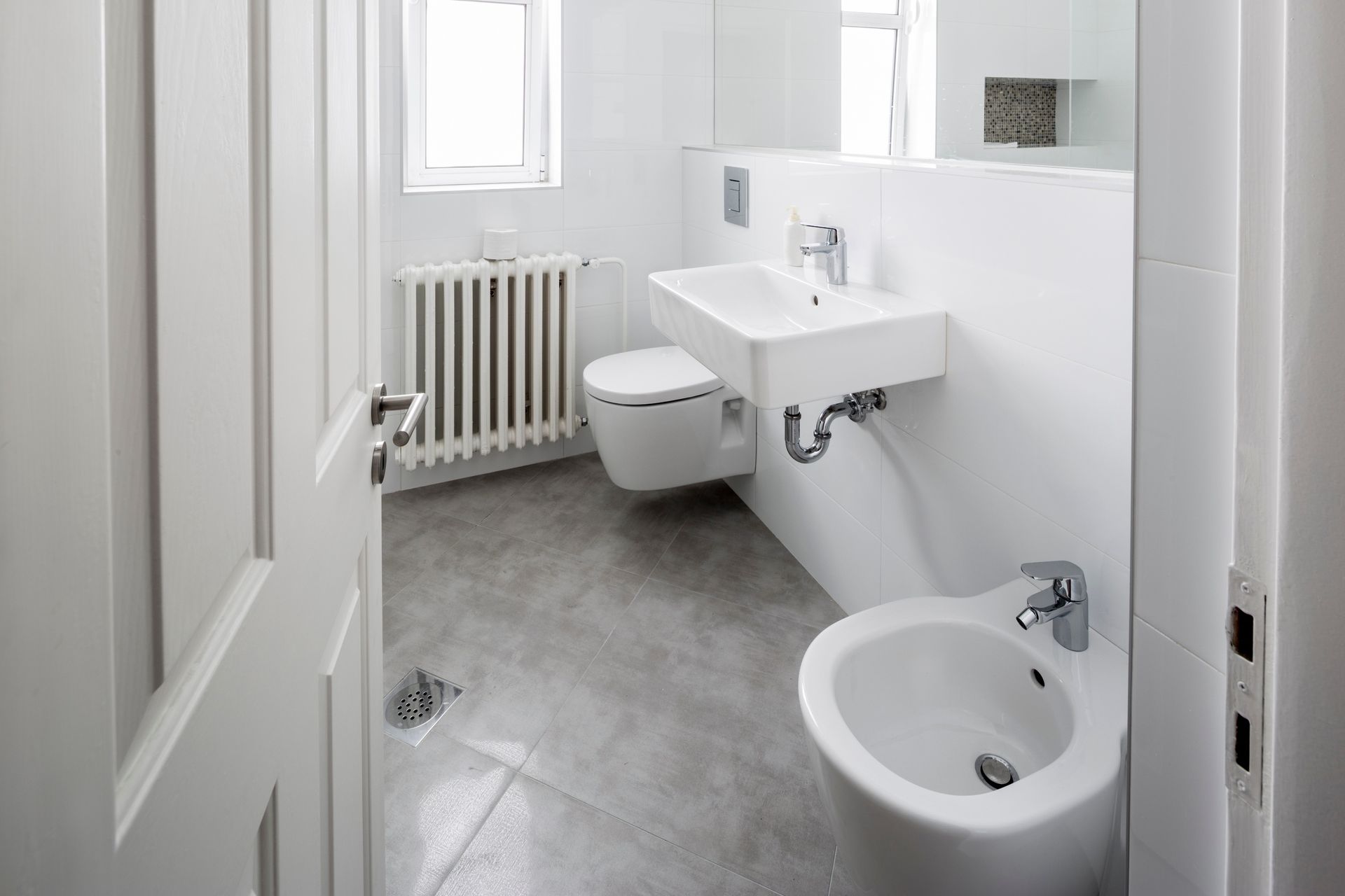 Small white bathroom with sink, toilet, bidet, radiator, and tiled floor near a window.
