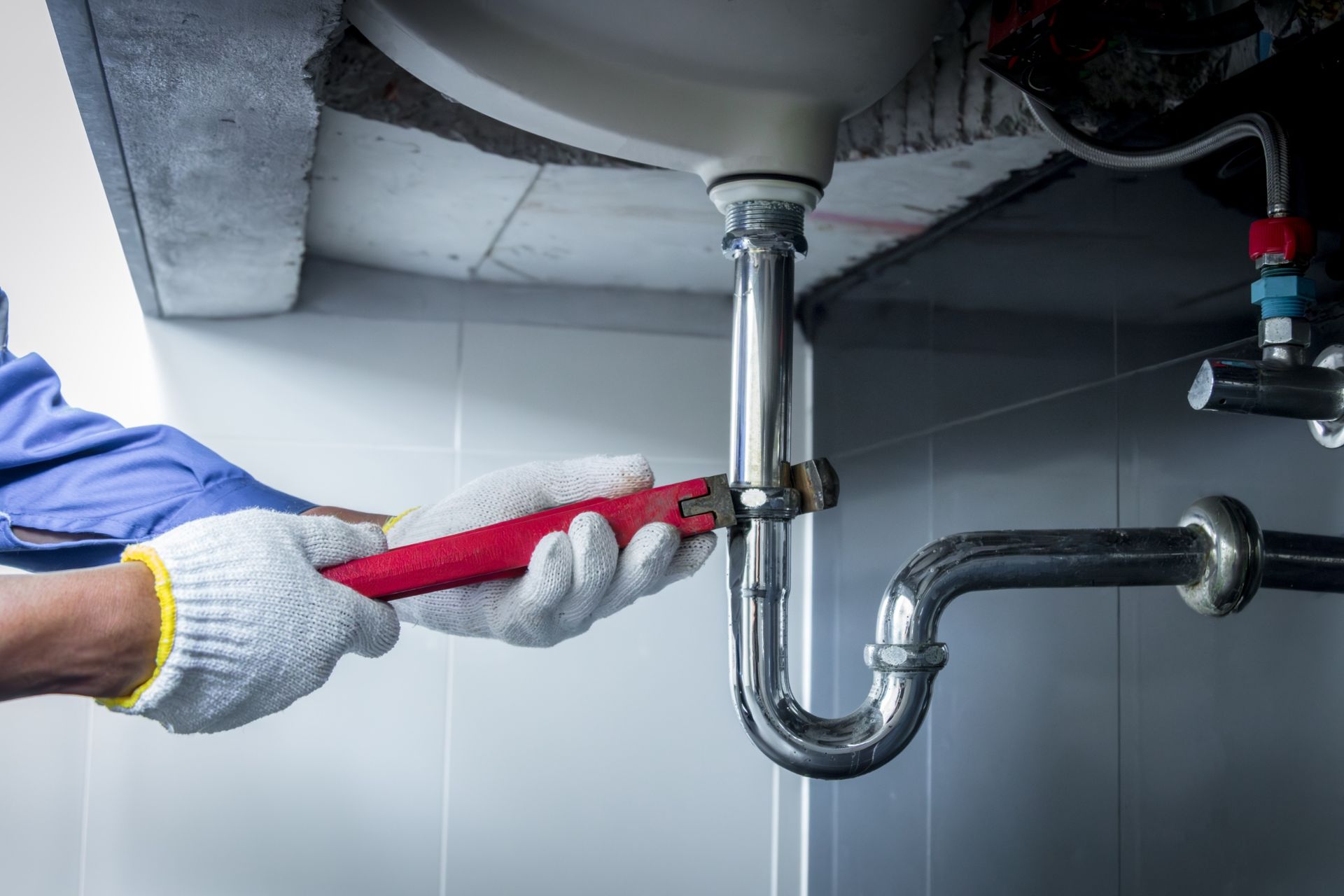 Gloved hands use a red wrench to tighten a pipe under a sink