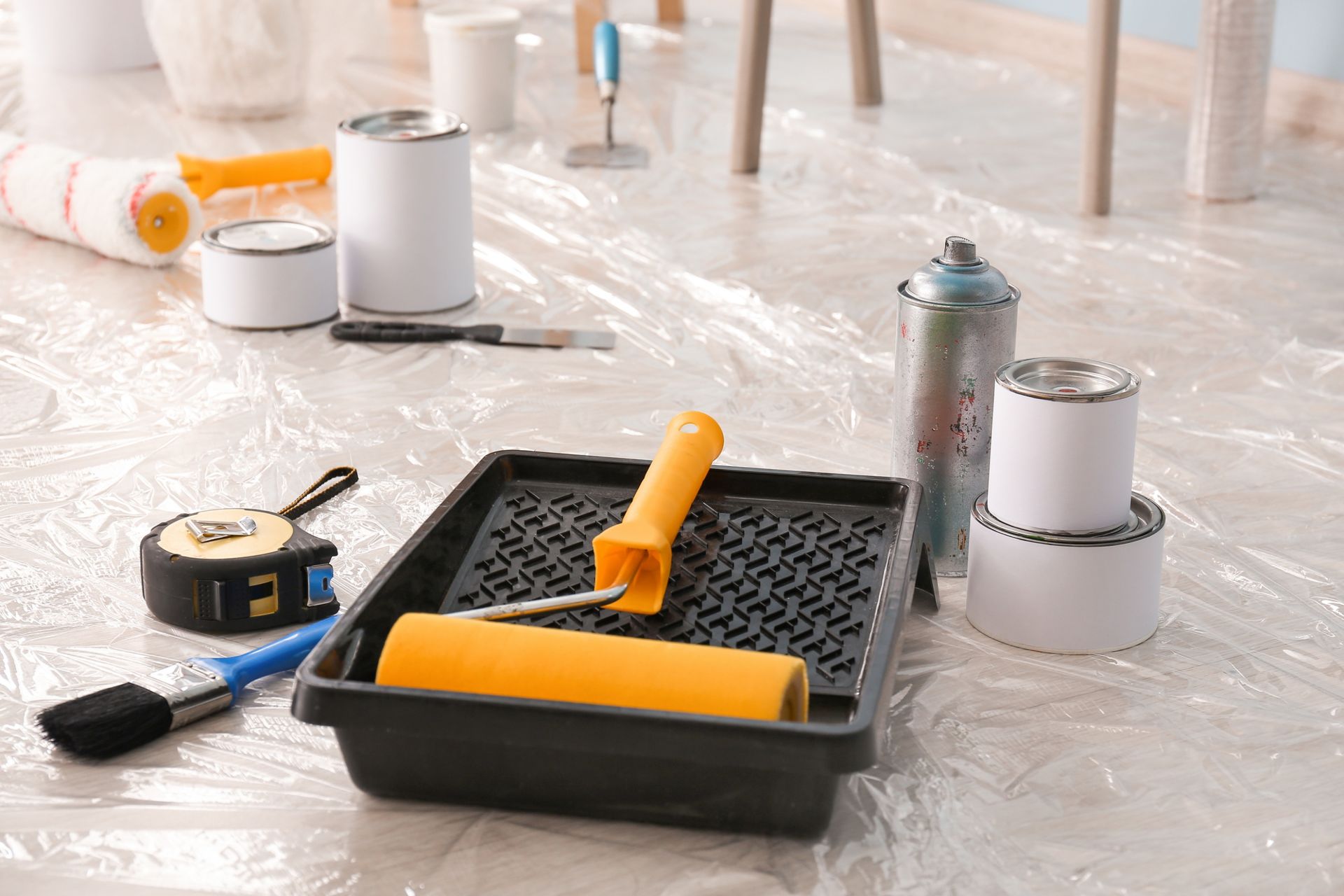 Painting supplies on a plastic-covered floor, including rollers, tape, and spray cans.