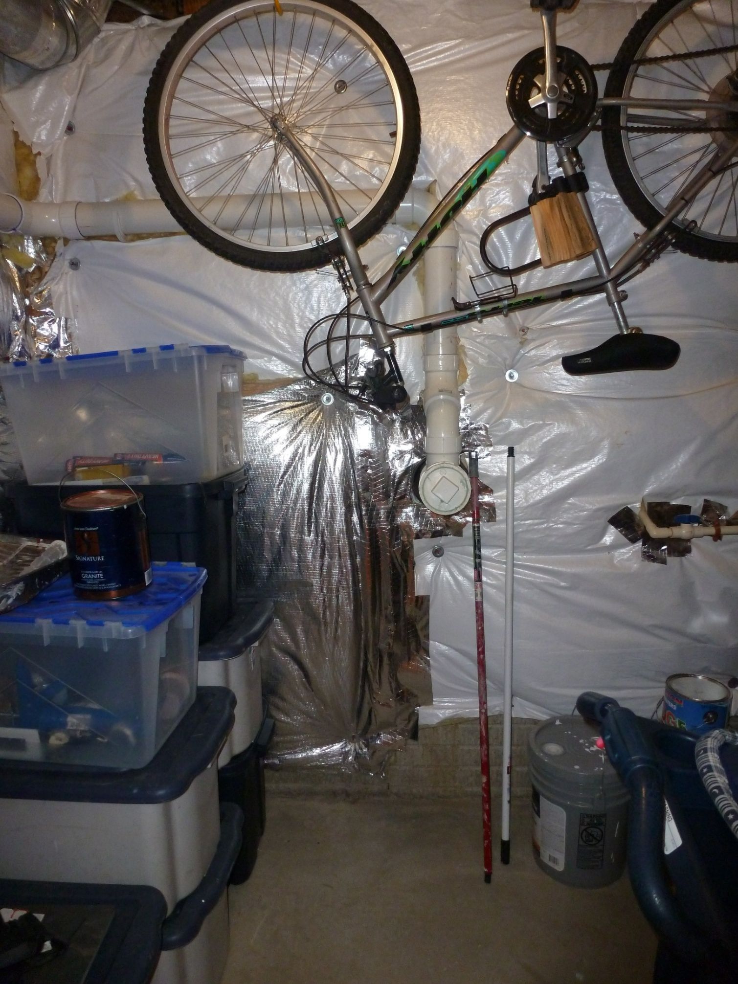 Cluttered basement with hanging bicycles, plastic storage bins, and a tripod stand against plastic-covered walls