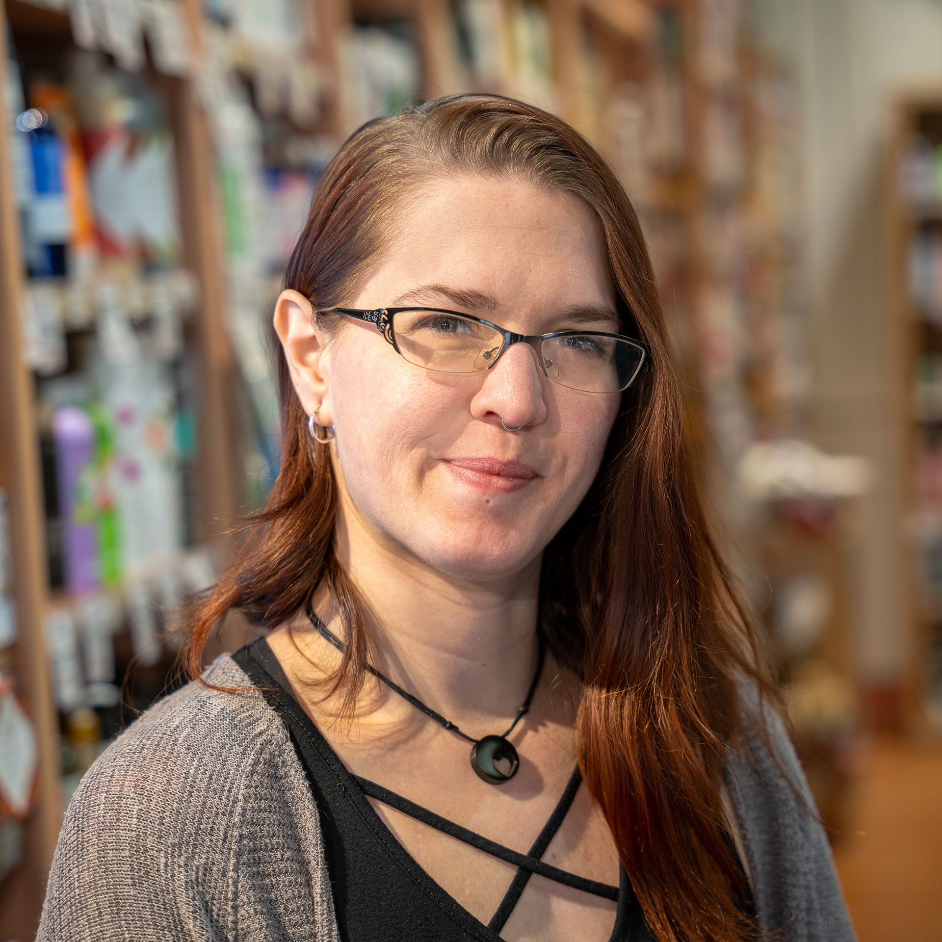 A woman wearing glasses and a necklace is standing in front of a bookshelf.
