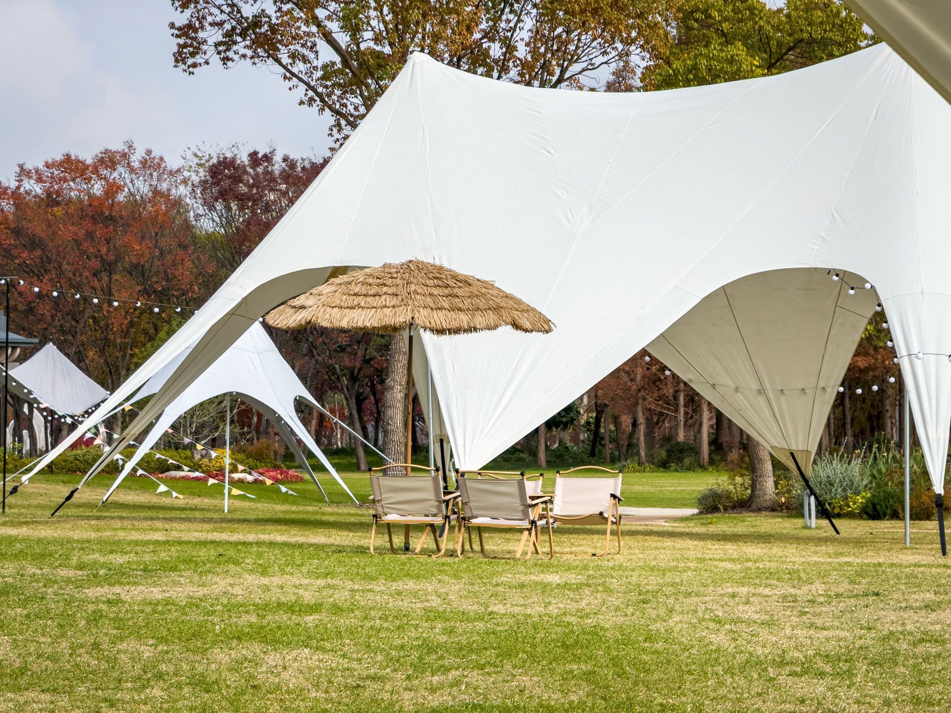 White tensile canopy shelters a patio with chairs and umbrella in a grassy park setting