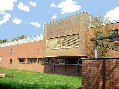 Ein Backsteingebäude mit großen Fenstern und einem Glasvordach, neben einer Rasenfläche unter blauem Himmel.