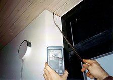 Eine Hand hält ein digitales Messgerät in der Nähe einer Deckenecke mit Kabeln und einem Fenster und misst oder prüft.
