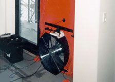 Ein schwarzer Ventilator ist an einer orangefarbenen Wand neben einer Tür montiert, die Kabel liegen auf dem Boden.