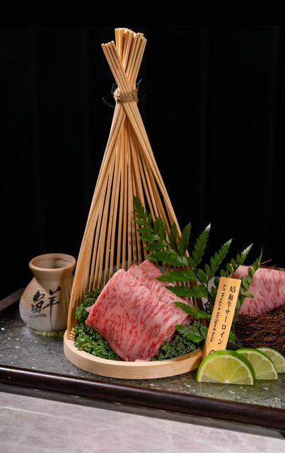 Sliced wagyu beef on a platter with greenery, lime wedges, and a small sake cup on a dark table.