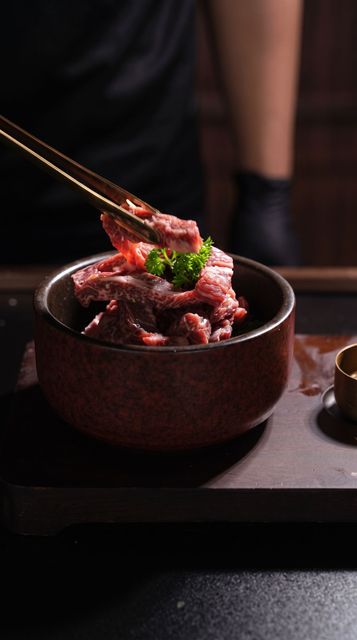 Chopsticks lifting glazed meat from a dark bowl, topped with green garnish, on a dimly lit table