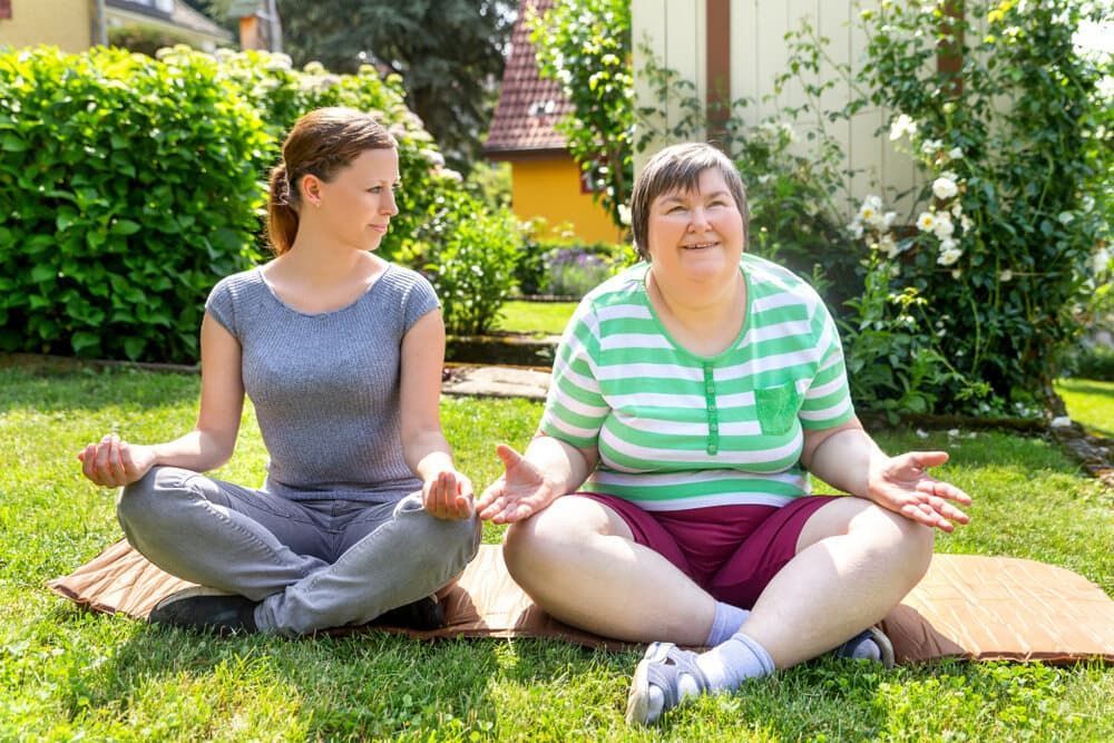 Two Women Are Sitting in a Lotus Position on a Yoga Mat in the Grass — Empowering You Disability Services In Maryborough, QLD