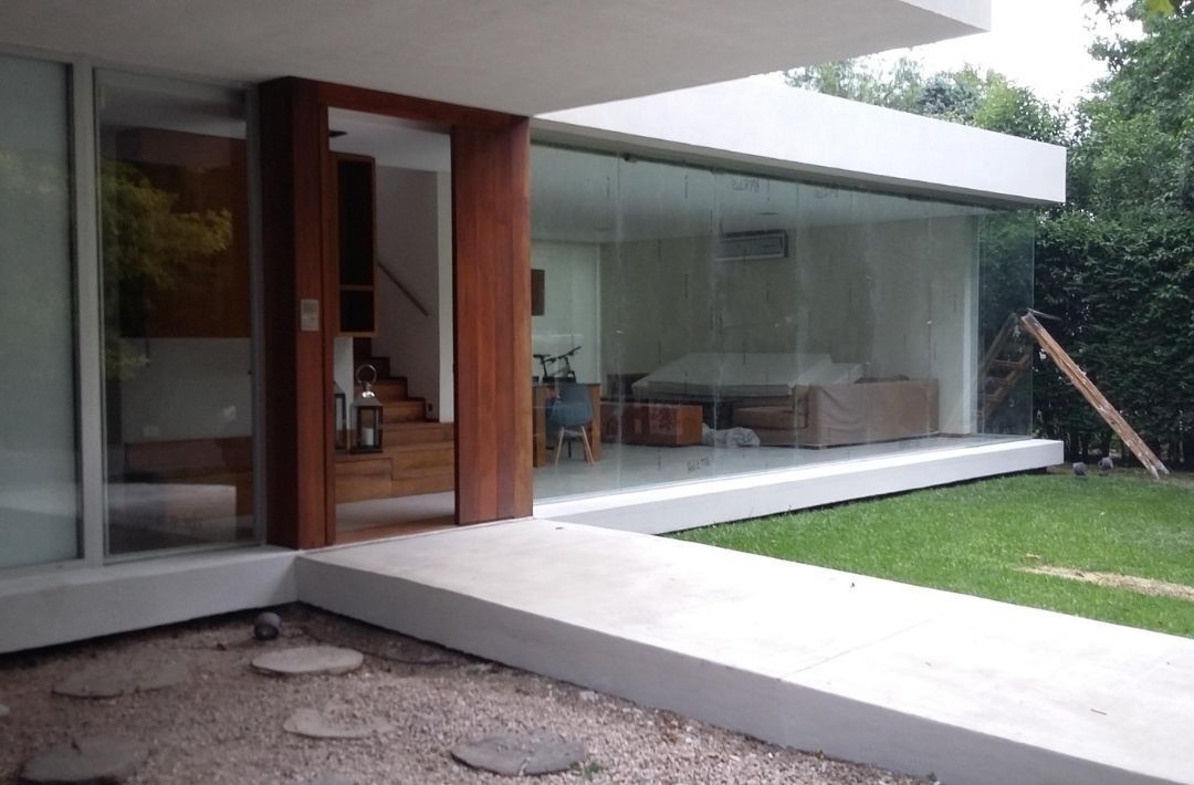 Patio de una casa moderna con grandes paredes de vidrio, entrada de madera y césped junto a un camino de concreto.