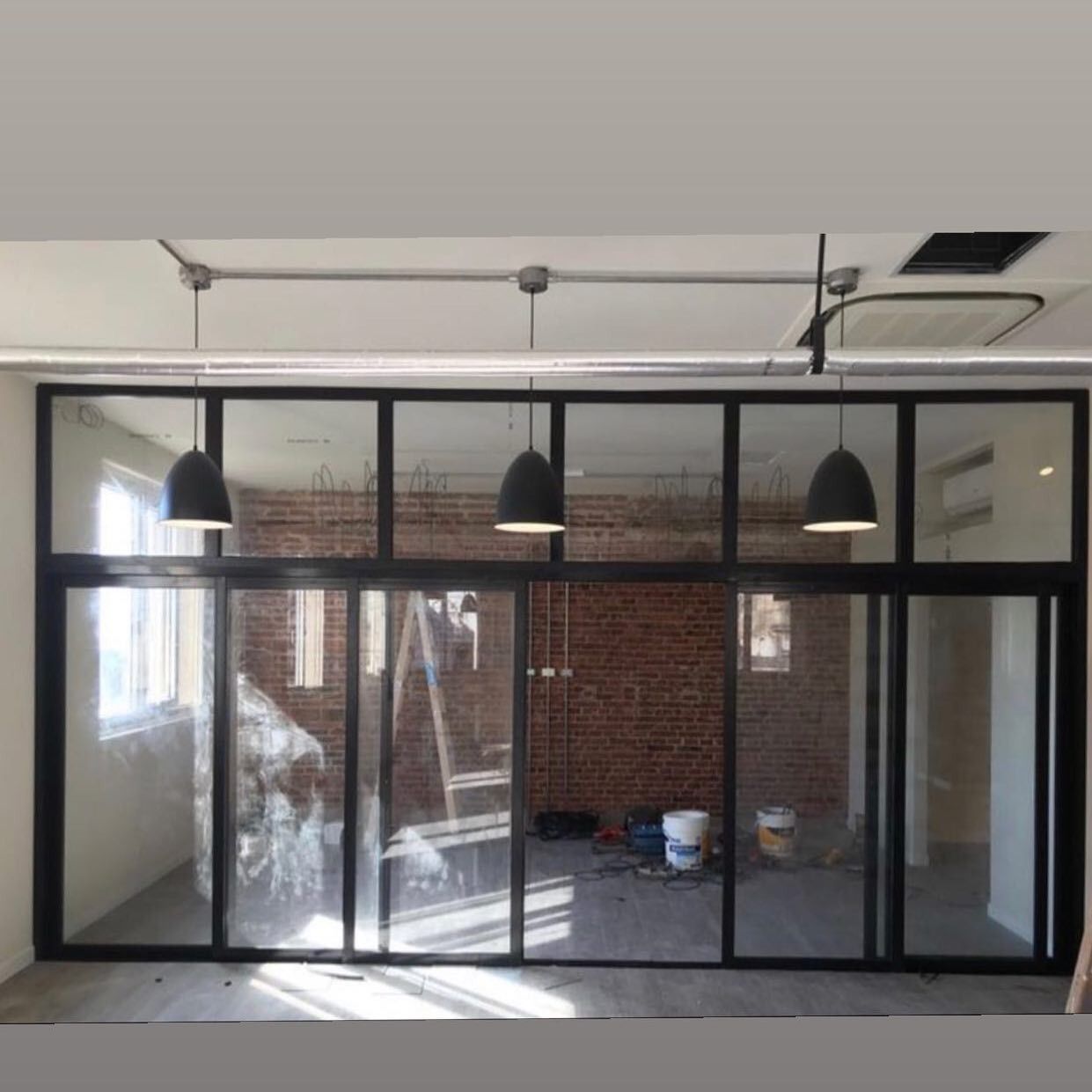 Oficina en construcción con paredes de cristal, lámparas colgantes negras y pared de ladrillo visto.