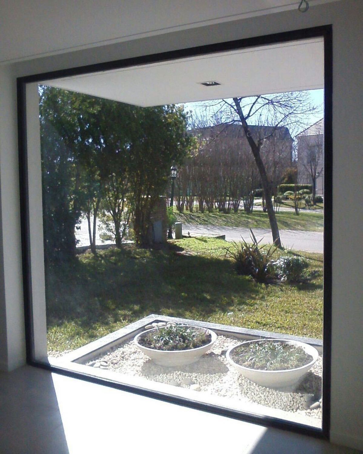 Vista a través de una gran ventana de un patio soleado con árboles, césped y dos macetas redondas blancas.