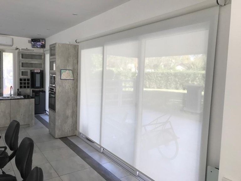 Salón moderno con persianas enrollables blancas, suelo de baldosas grises y sillas negras junto a una gran ventana.