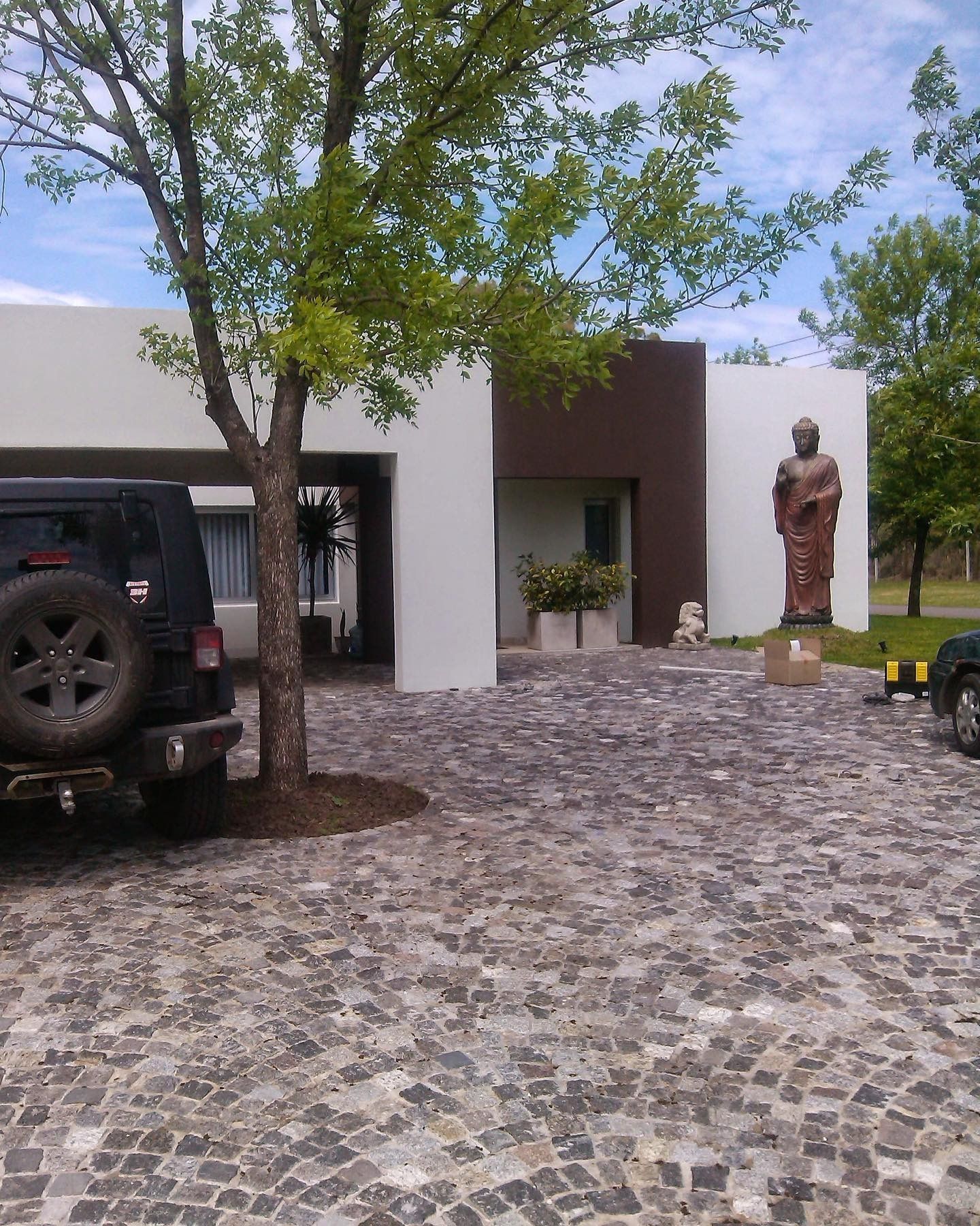 Entrada a un edificio moderno con camino empedrado, árboles, vehículos estacionados y una estatua junto a la puerta.