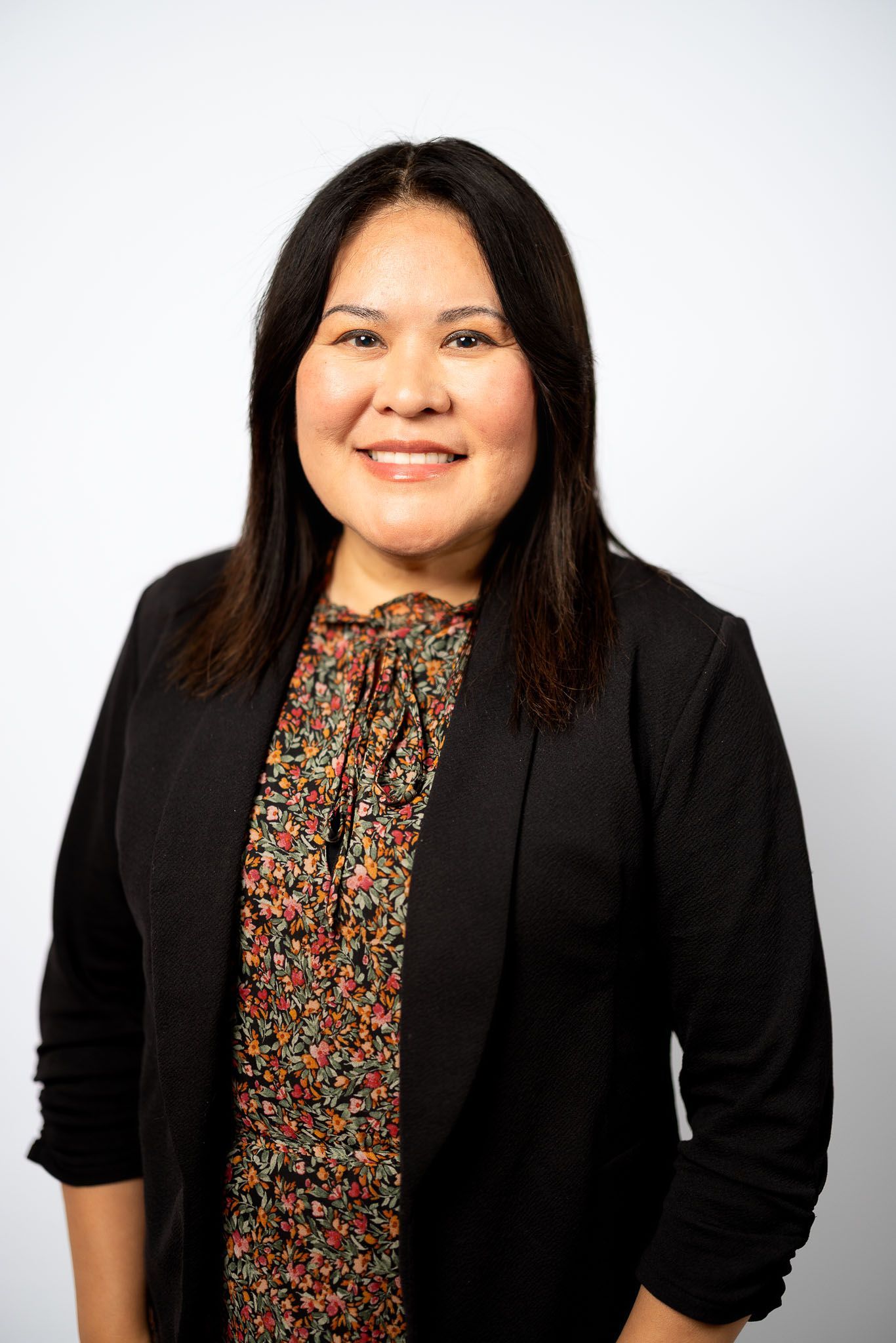 Woman smiling, wearing black blazer and patterned top, standing against a white backdrop.