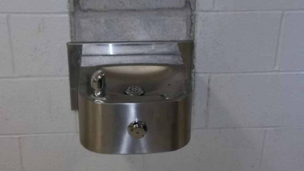 A stainless steel water fountain is mounted to a white brick wall.