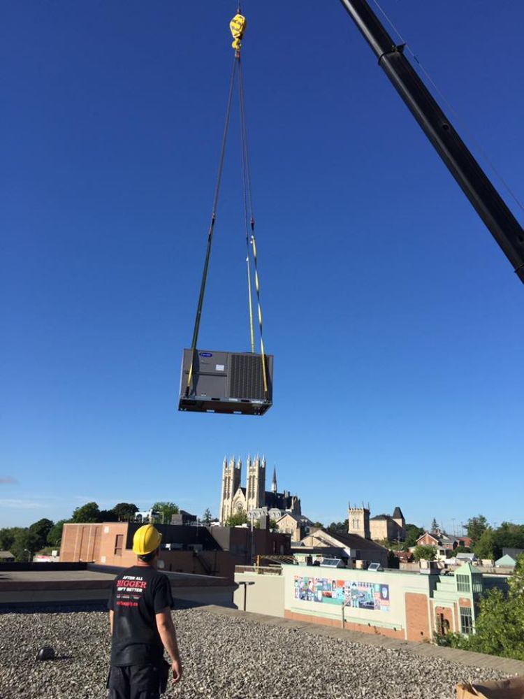 A man wearing a hard hat is standing on a roof watching a crane lift a machine into the air.