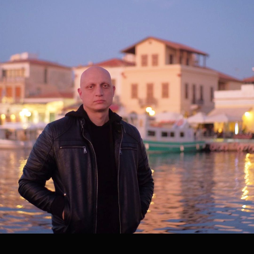 Άντρας με ξυρισμένο κεφάλι με μαύρο μπουφάν στέκεται δίπλα σε ένα λιμάνι με κτίρια και βάρκες το σούρουπο.