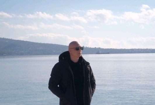 Άντρας με γυαλιά ηλίου και μαύρο μπουφάν στέκεται δίπλα στο νερό, κοιτάζοντας προς ένα βουνό στο βάθος.