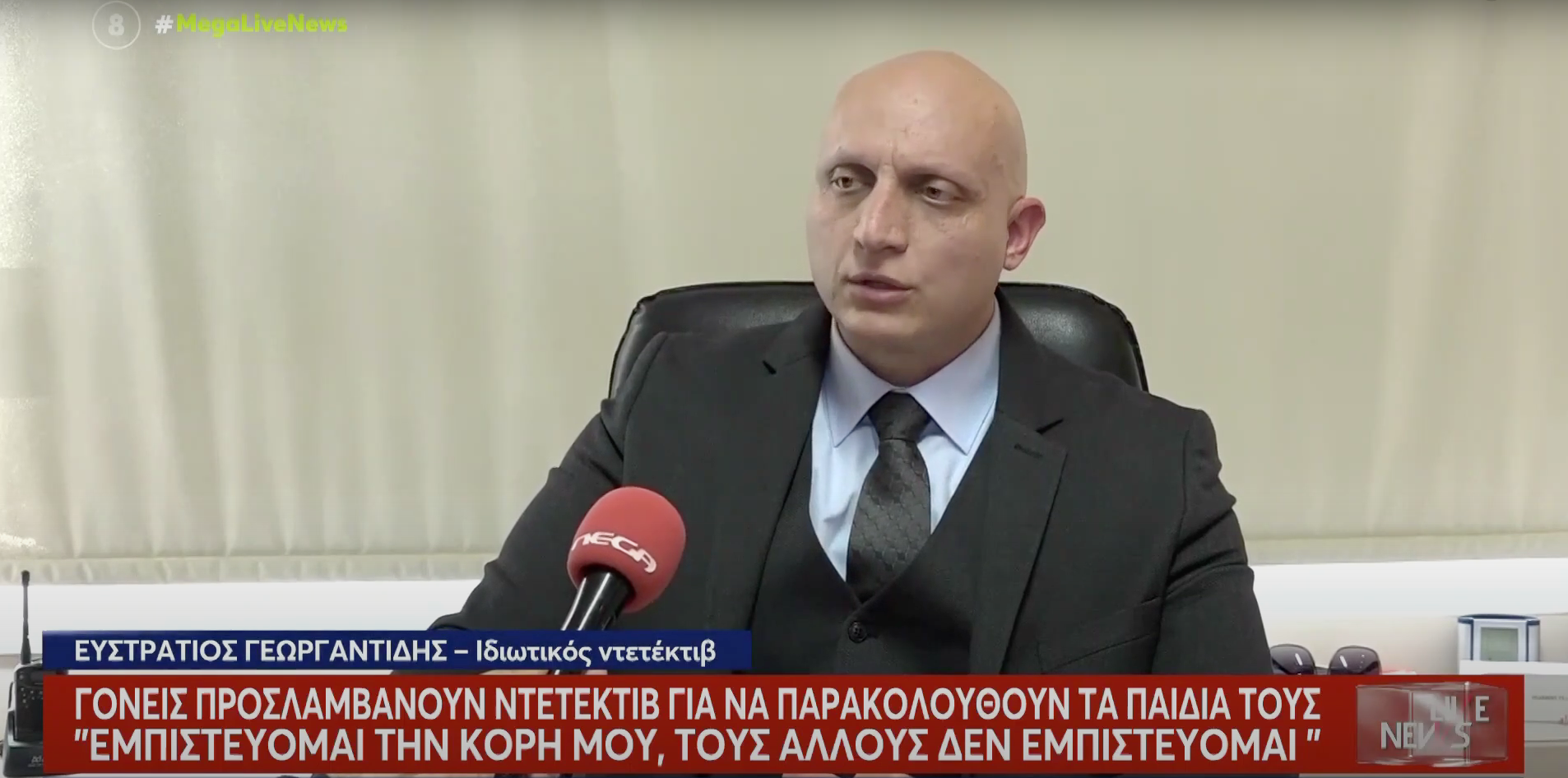 Άνδρας με κοστούμι μιλάει σε μικρόφωνο, με τίτλο είδησης στα ελληνικά.